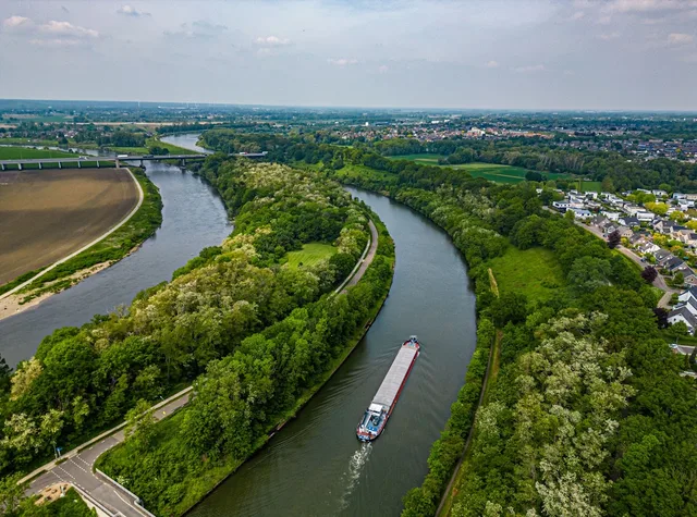 Aerial of a cargo barge working the river Maas between green banks, town on the horizon