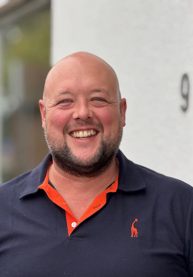 Bald man with a beard in a navy polo laughing straight into the camera