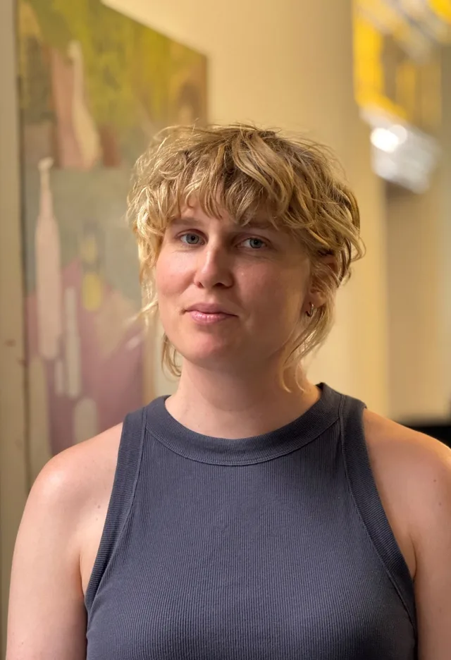 Calm portrait of a person with curly blonde hair in a grey tank top in front of a painted wall