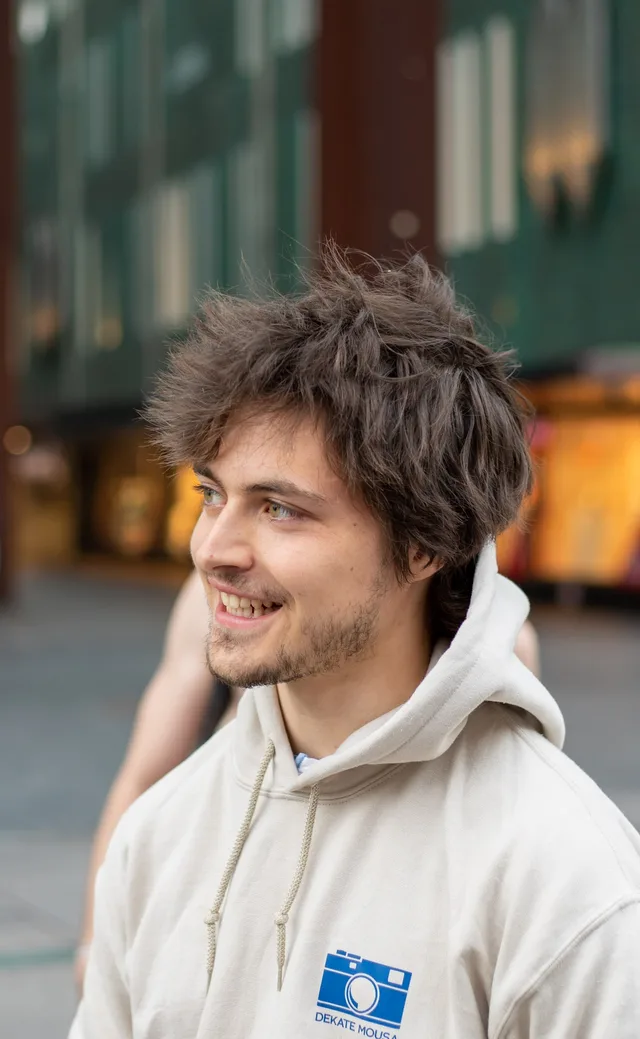 Young man in a cream hoodie glancing sideways with a smile on a shopping street