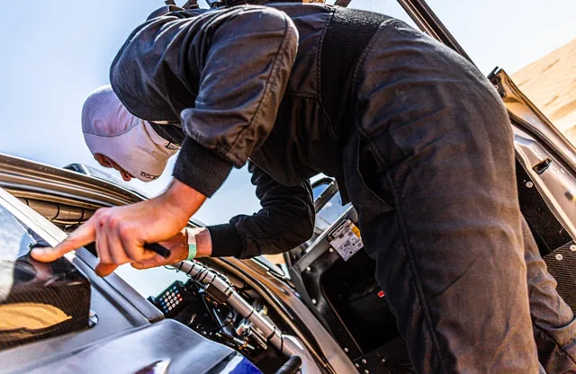Crew member in black racing overalls leaning into a rally car cockpit during a desert stage