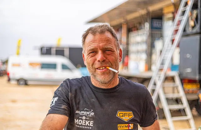 Rally mechanic with a cigarette between his lips grinning at the camera in front of the service trucks