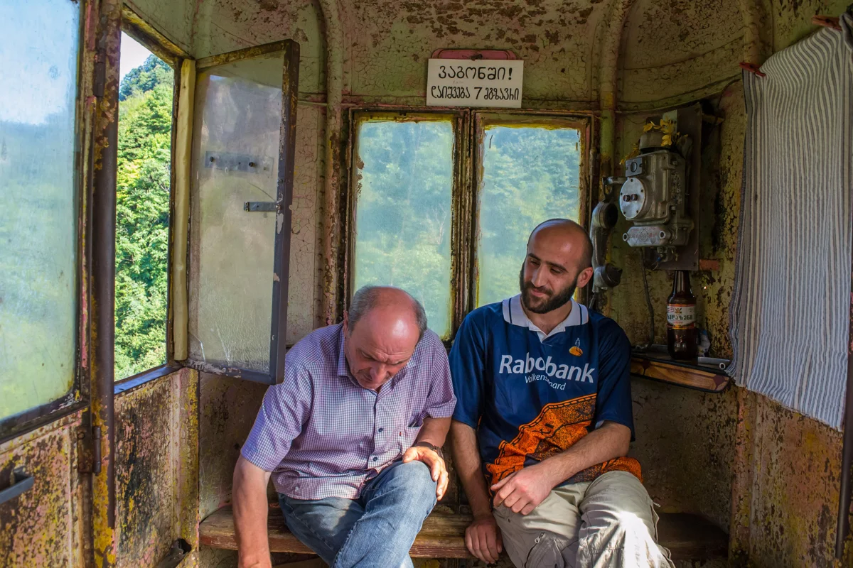 Two men inside a rusty Soviet-era cable car cabin in Georgia, forested hills through the windows